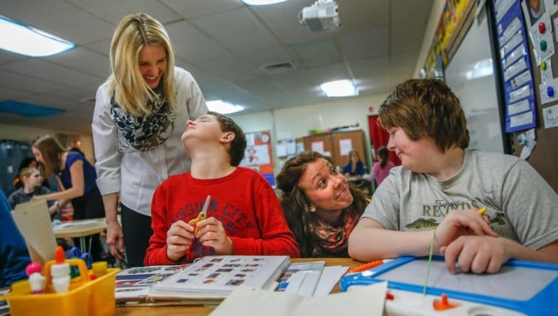 Two teachers laugh and smile with two students sitting at a desk in a classroom.
