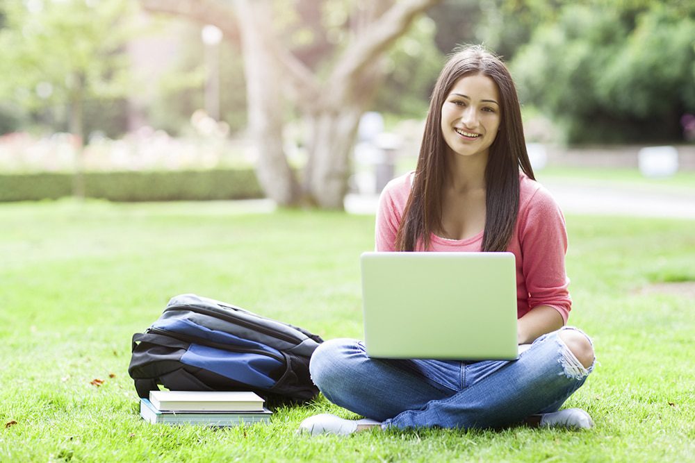 A girl sitting crosslegged outdoors smiles at the camera with a laptop in her lap and a backpack next to her on the ground.