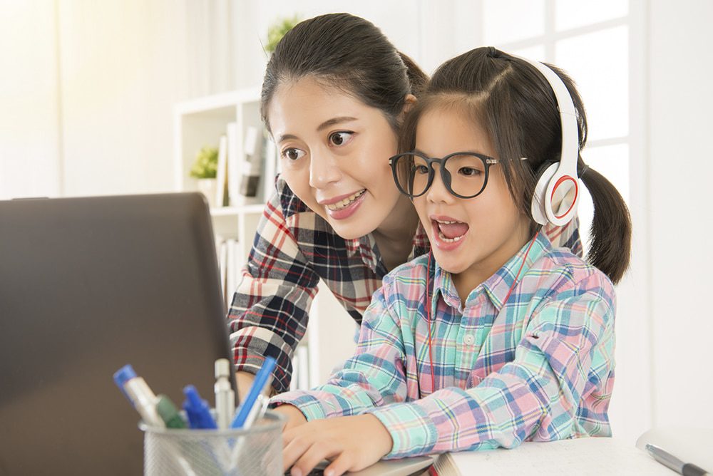 A mother smiling with her daughter who is wearing headphones and glasses while working at a computer in a home