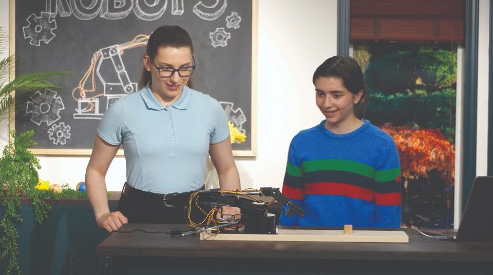 2 students look animatedly at a incomplete robot in front of them on a table.