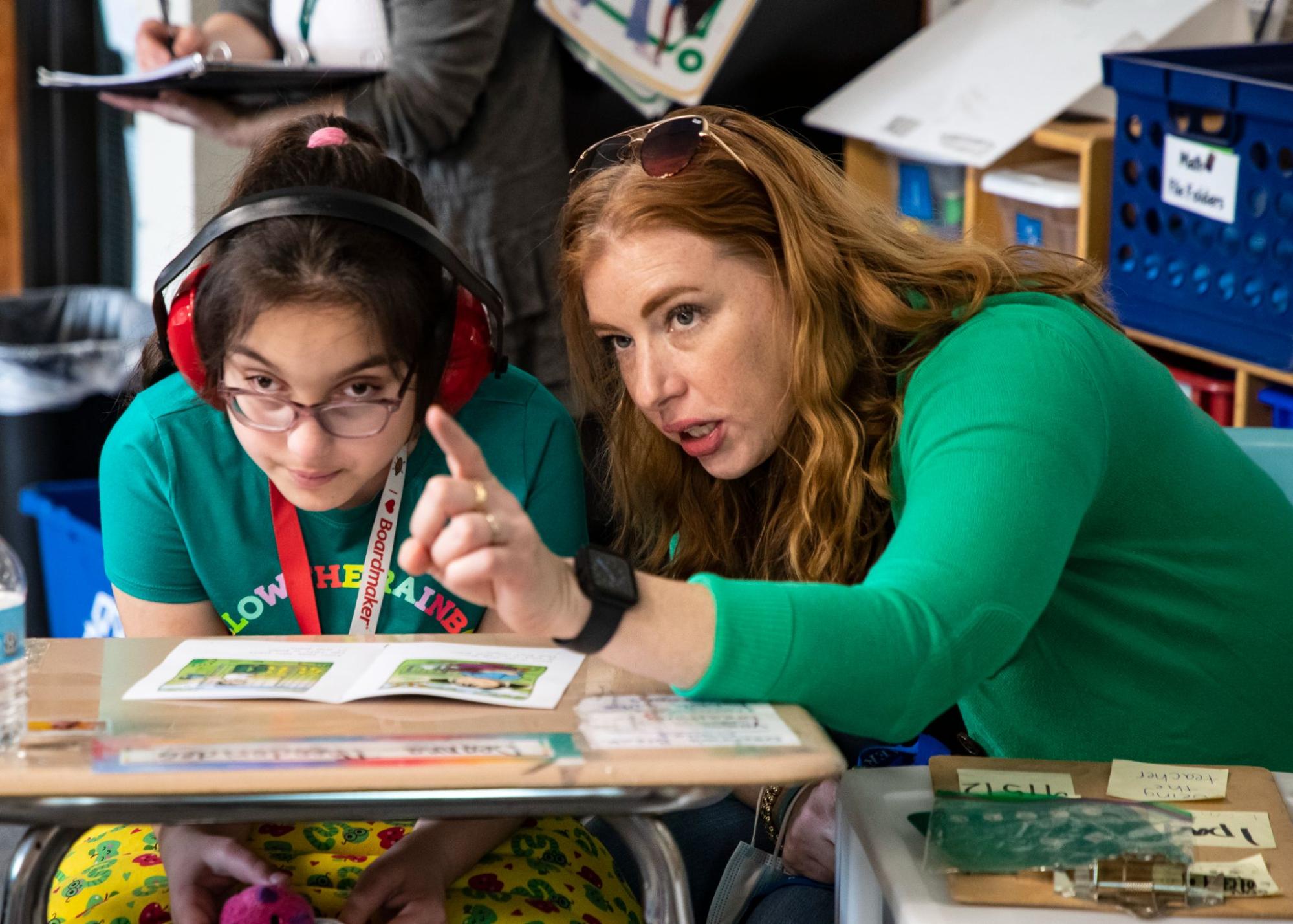 In a classroom, a teacher wearing a green shirt, points something while working with a student sitting at a desk.