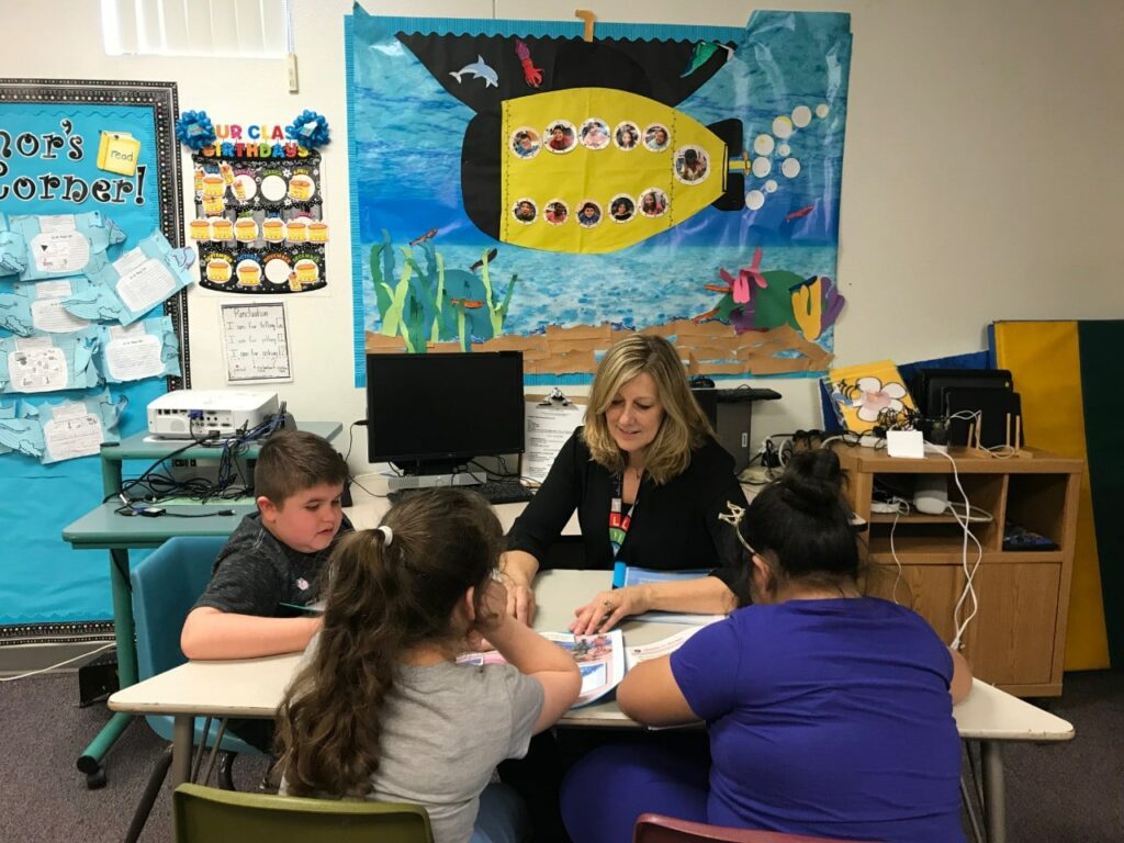 Teacher reads at a desk with three children in a classroom
