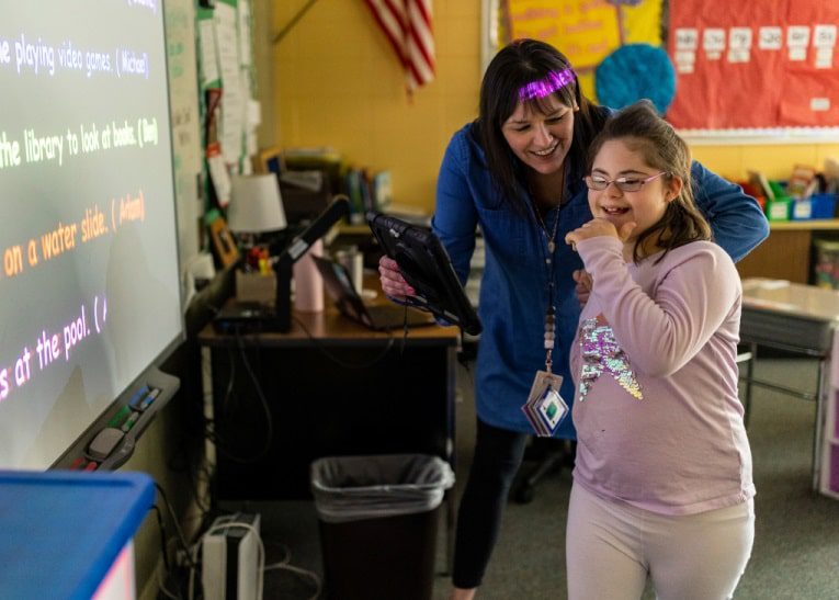 A teacher smiles at a laughing student, while the student presents in front of a smart board in a classroom.