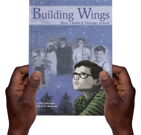 2 hands holding the "Building Wings: How I Made it through School" book, with an animated picture of Don Johnston on the cover.
