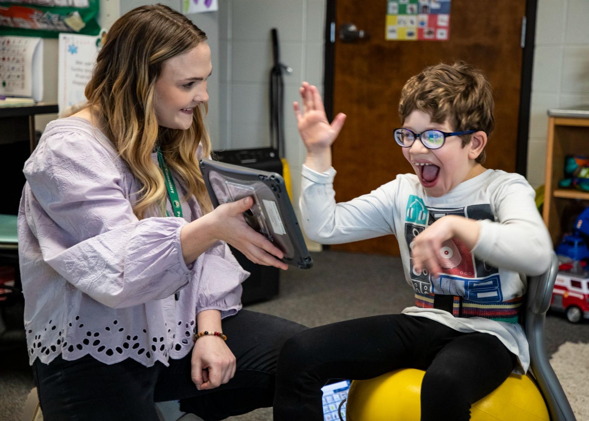 In a classroom, a teacher holds up an AAC device to a student who is excited and smiling.